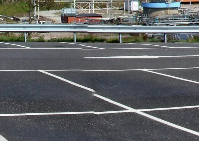 Stitched panorama of a paved yard with a visible vertical seam where painted lines and a guard rail break and shift sideways