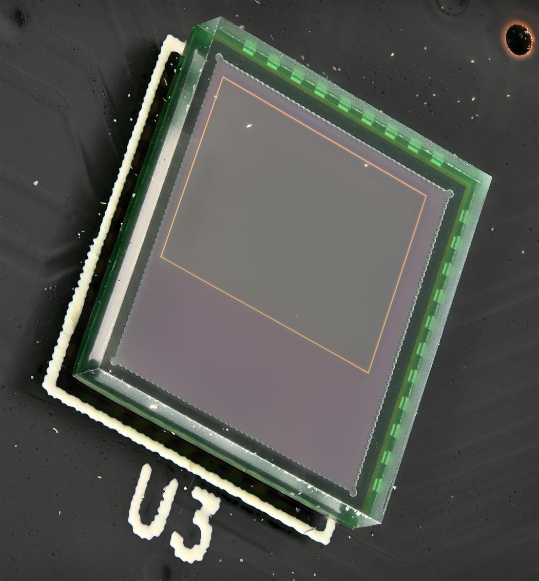 Macro photograph of a CMOS image sensor in a glass package, the dark active imaging area framed by a thin gold line