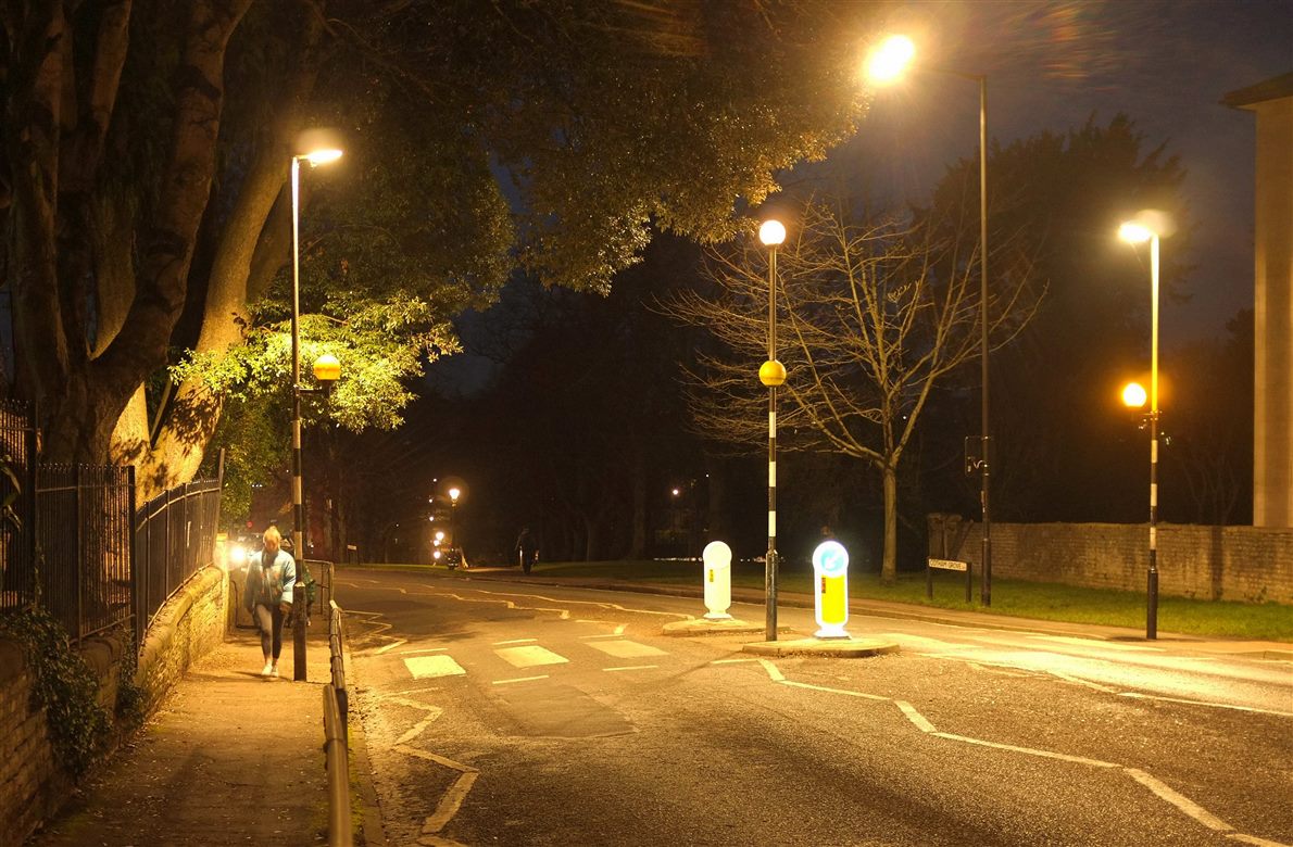 Night street scene lit by orange sodium lamps, a pedestrian on the pavement near an unmarked crossing