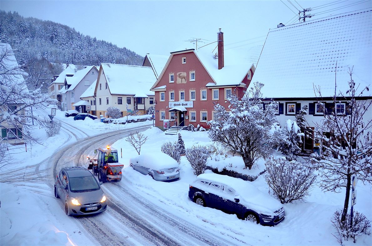 Snow-covered street with parked cars under snow and a vehicle driving on packed white road, markings buried