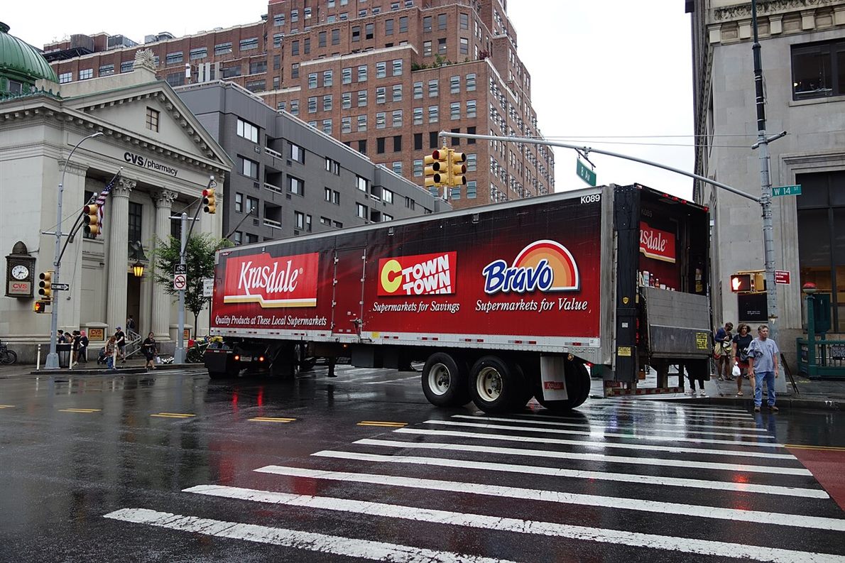 An articulated truck turning across a city crosswalk on a wet street, the trailer cutting inside the cab's path with pedestrians waiting at the corner