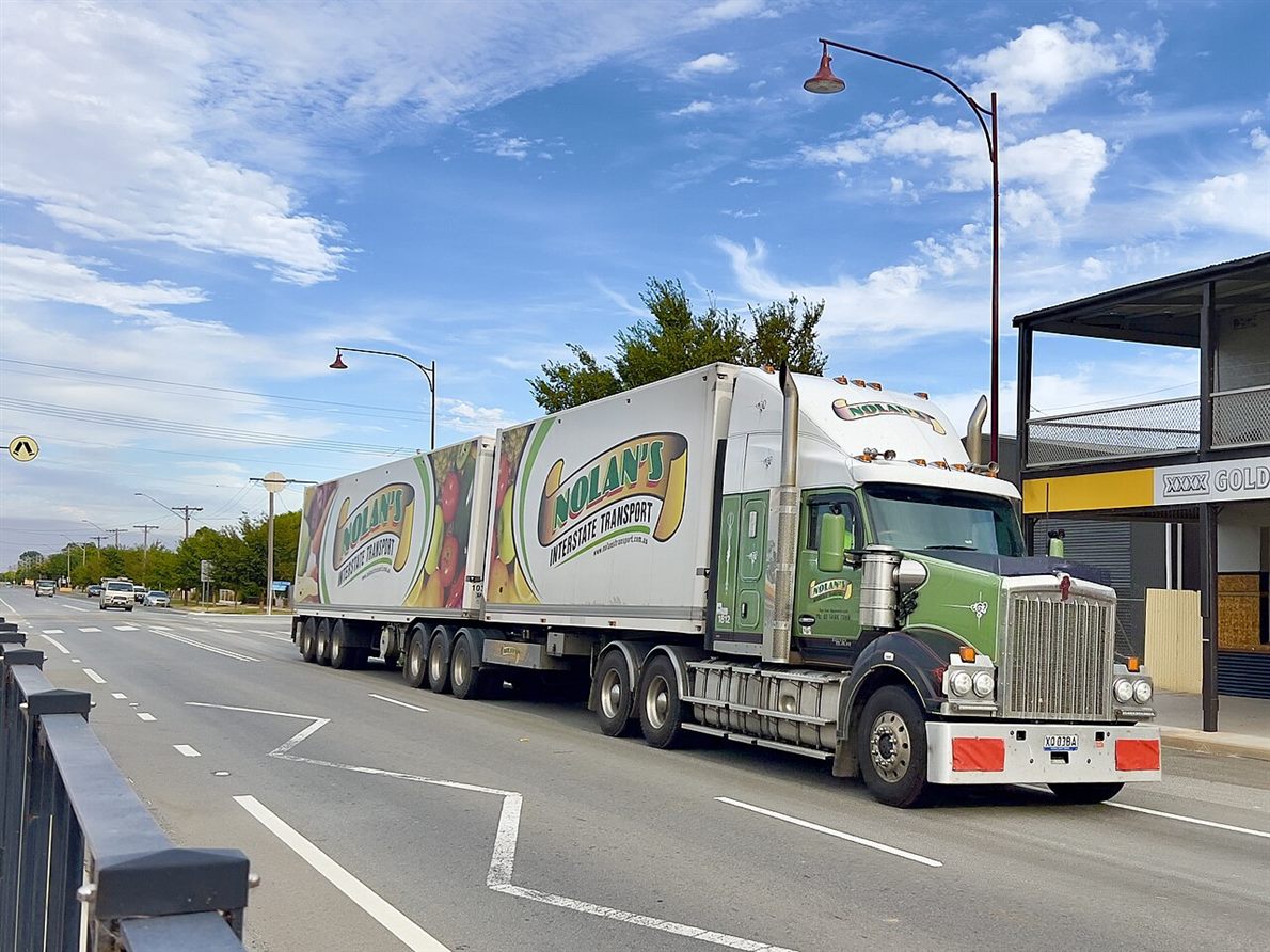 A B-double truck on a town street, a prime mover pulling two trailers coupled in line, seen from the side