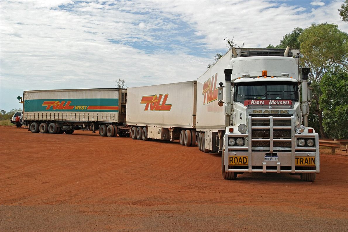 An Australian triple road train parked at a slight angle on red dirt, a prime mover and three trailers coupled in a line with the gaps and joints between them visible