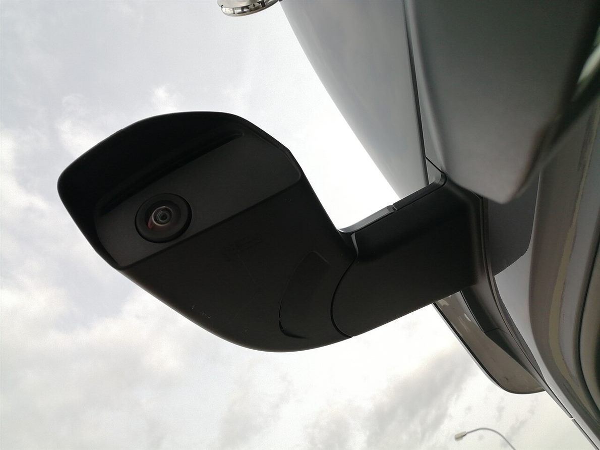 Close view of a camera unit mounted on a truck cab arm, the small lens visible in its housing against a cloudy sky