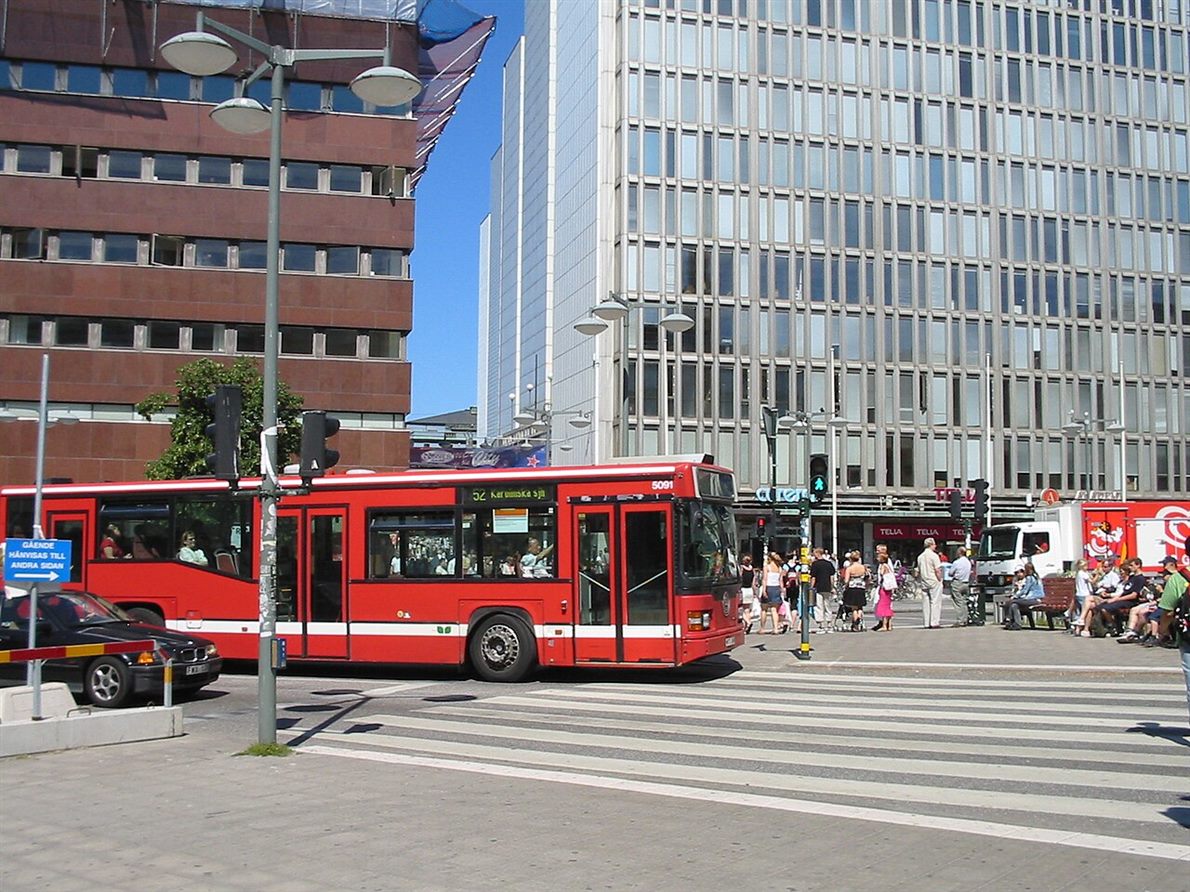 A red city bus turning at an intersection with pedestrians waiting on the corner and crossing the marked crosswalk
