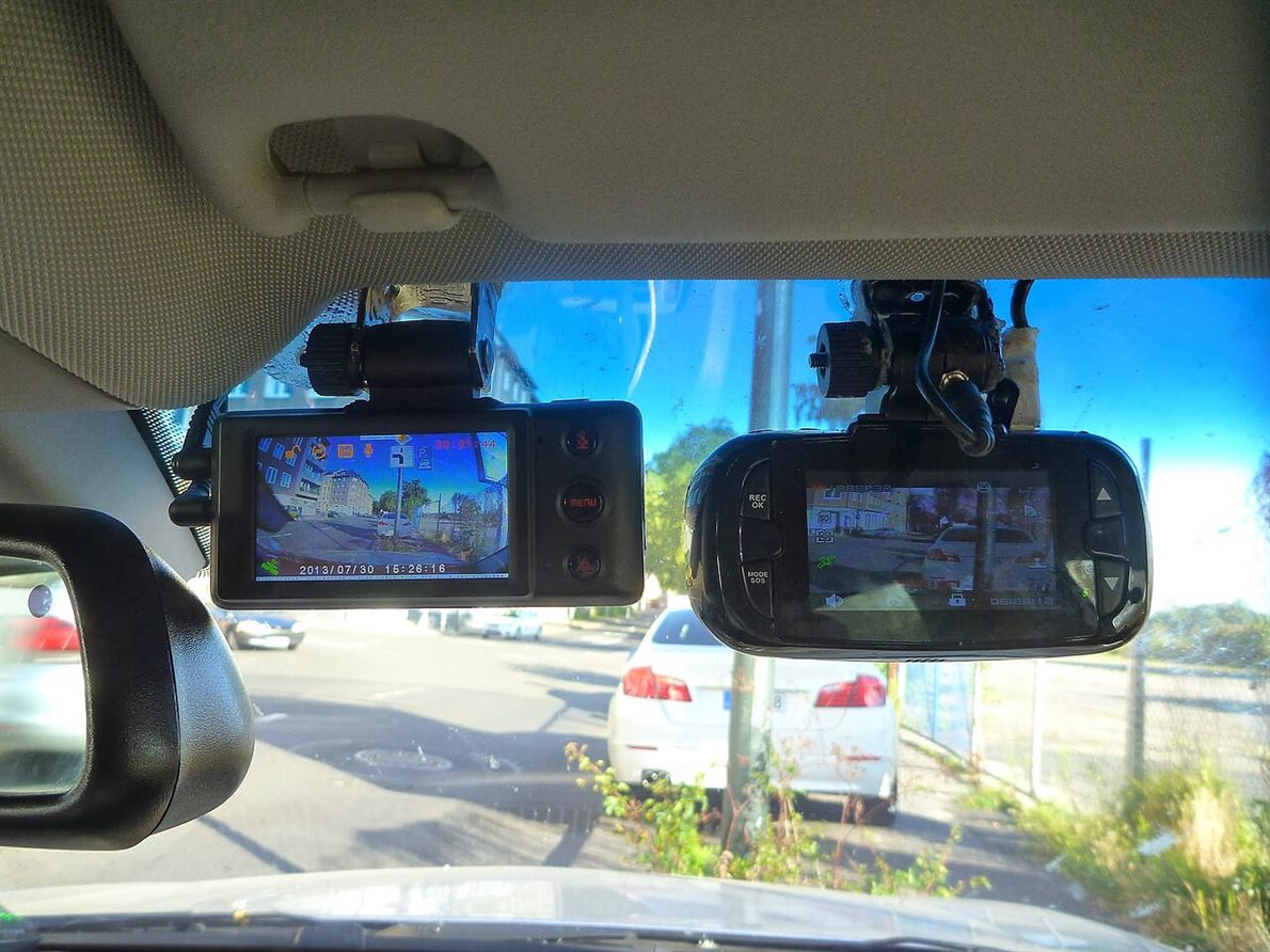 Two forward-facing cameras mounted on a vehicle windscreen, their screens showing the road ahead, parked cars visible through the glass