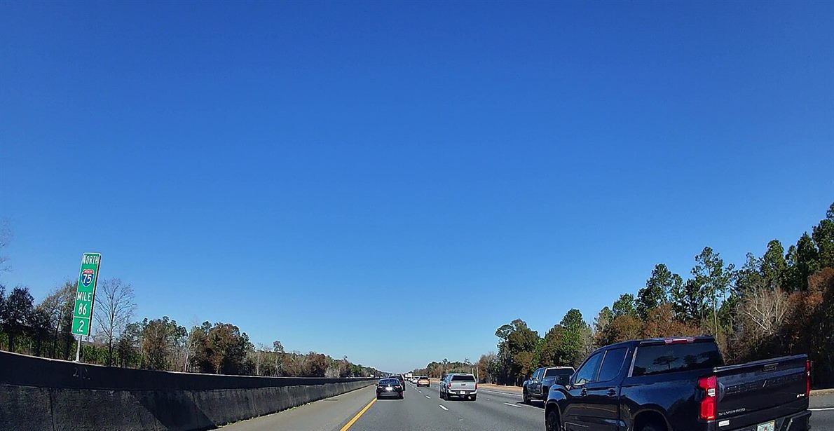 A forward view down a multi-lane highway from a vehicle, with cars and a pickup truck ahead in the lanes under a clear sky