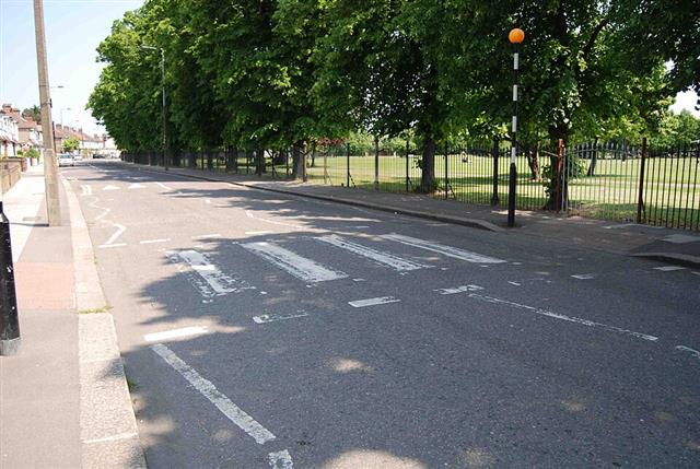 A suburban road with a badly faded pedestrian crossing and worn lane markings, dappled with shadows from roadside trees
