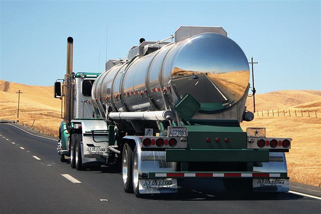 A chrome tanker semi-truck driving along a two-lane highway, holding its position within the lane between the dashed centerline and the solid white edge line