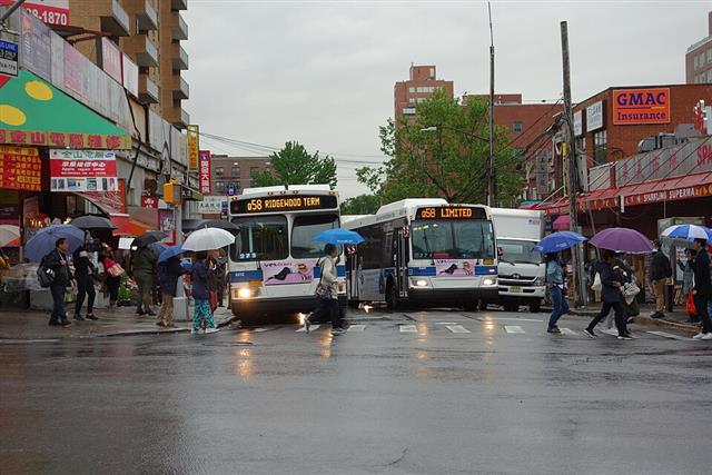 Two city buses at a busy wet urban intersection with many pedestrians carrying umbrellas crossing directly in front of them