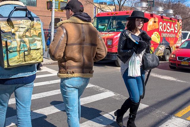 Three pedestrians in varied winter clothing crossing a sunny city street on a marked crosswalk, partly overlapping, with a red food truck and a car behind them