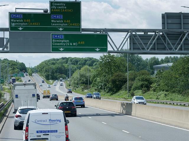 Driver's view along a motorway toward two overhead green sign gantries and a steel sign truss, with vans and cars in the lanes ahead