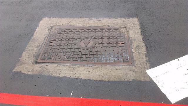 A rectangular cast-iron manhole cover set flush in an asphalt road surface beside painted road markings
