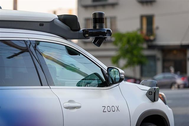 A white autonomous test vehicle with a roof-mounted sensor pod holding a spinning laser scanner and several cameras, ZOOX branding on the door