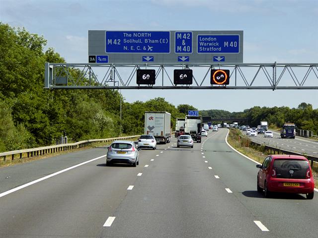 A steel sign gantry spanning a motorway, carrying direction boards and overhead speed signals, with cars and a lorry passing beneath it