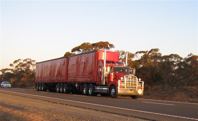 A red B-double truck pulling two trailers along a rural highway at dusk