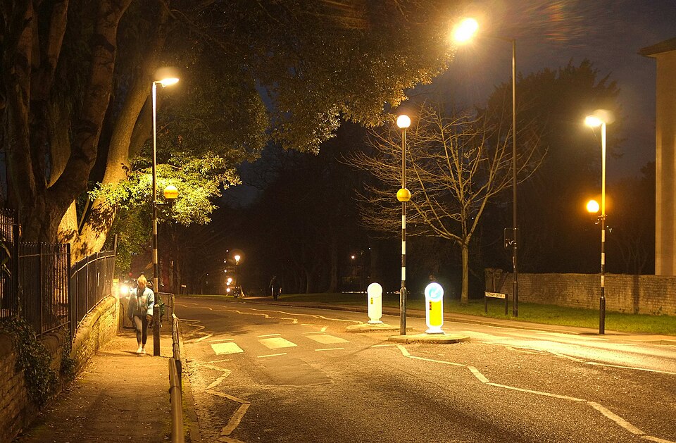 A pedestrian crossing at night, a person standing in shadow between pools of street light