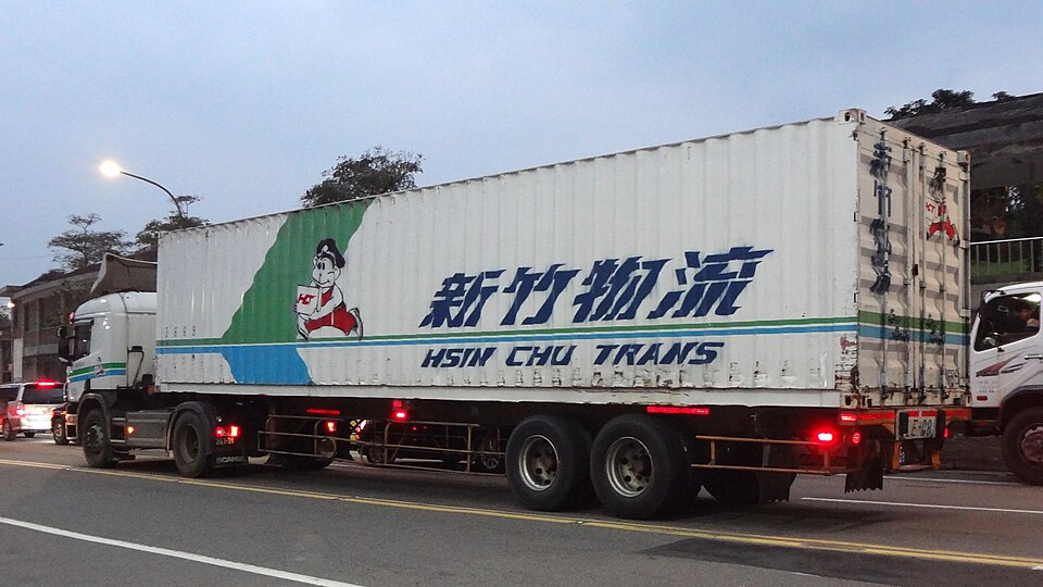 An articulated goods truck on a city road at dusk with its lights on