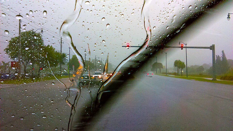 The view through a rain-covered windscreen with a wiper streak, a wet road and traffic lights ahead