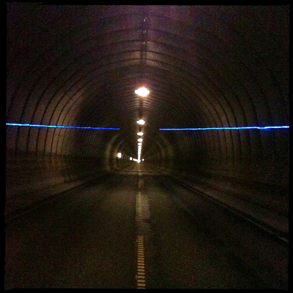 The dim interior of a road tunnel with lights along the roof and a small bright exit far ahead